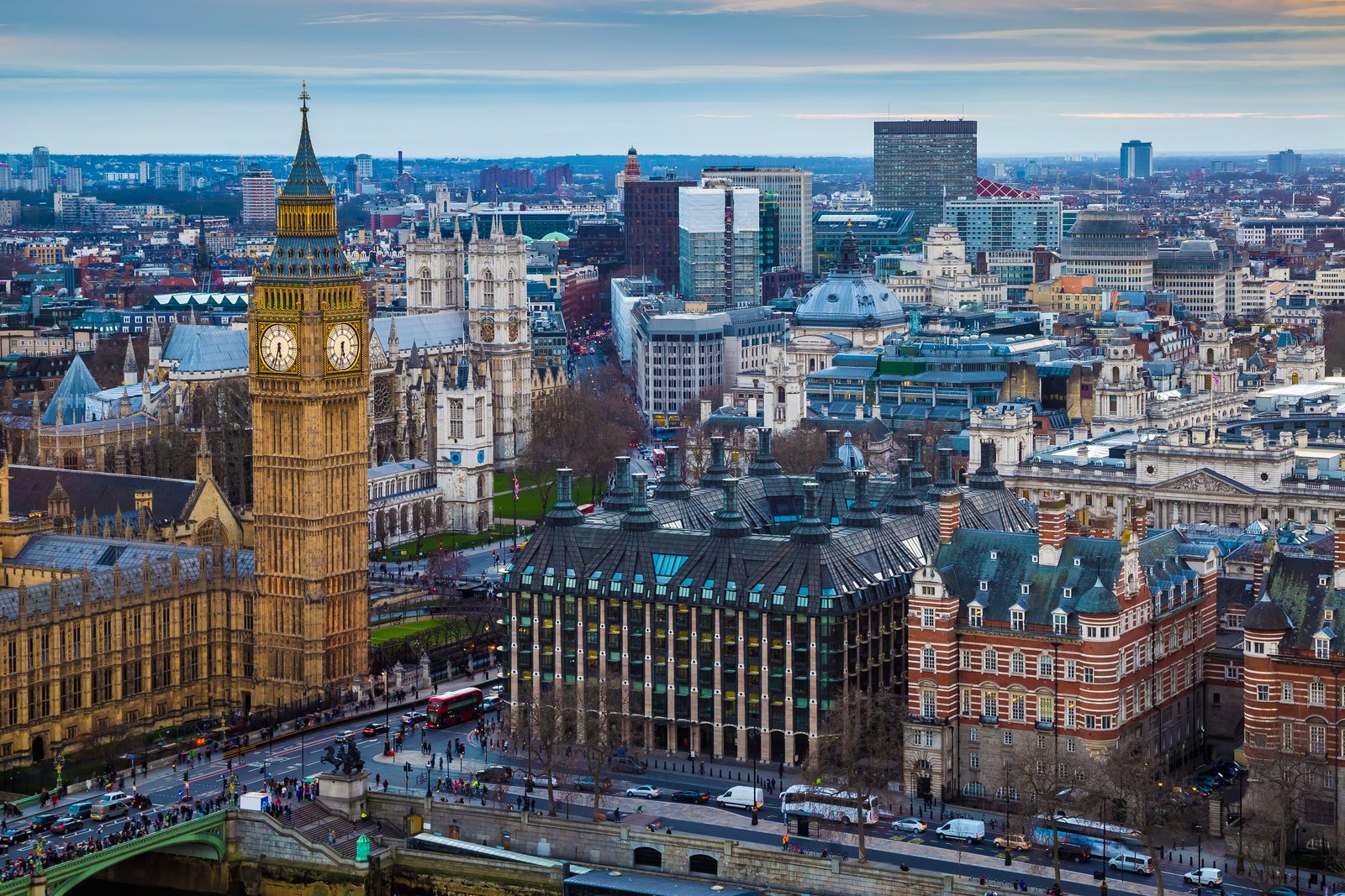 Aerial view of Big Ben and the Houses of Parliament with Westminster Abbey and the London skyline behind, a red double-decker bus crossing Westminster Bridge in the foreground